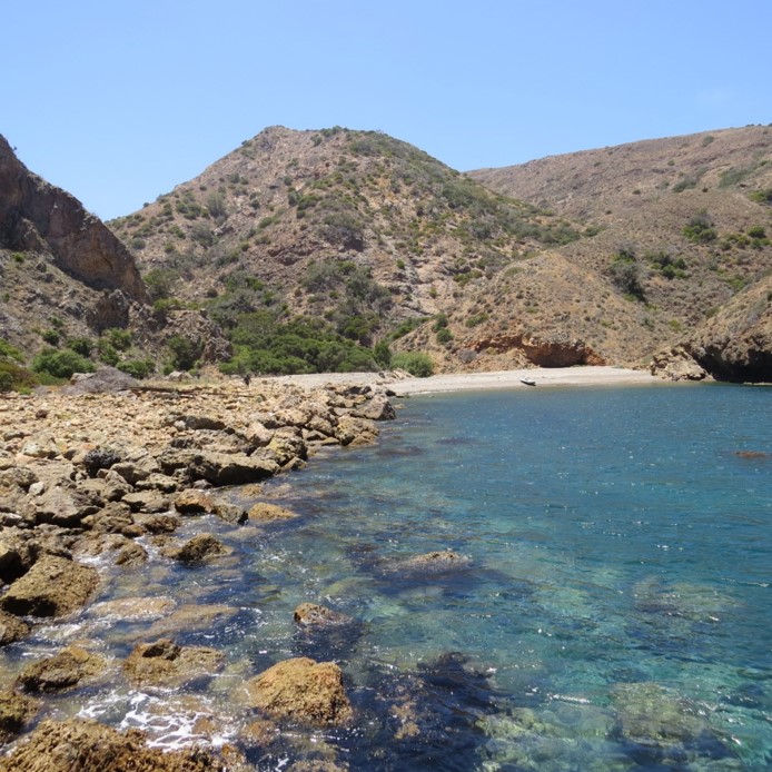 Rocky shoreline with clear blue water and hilly terrain under a clear sky.