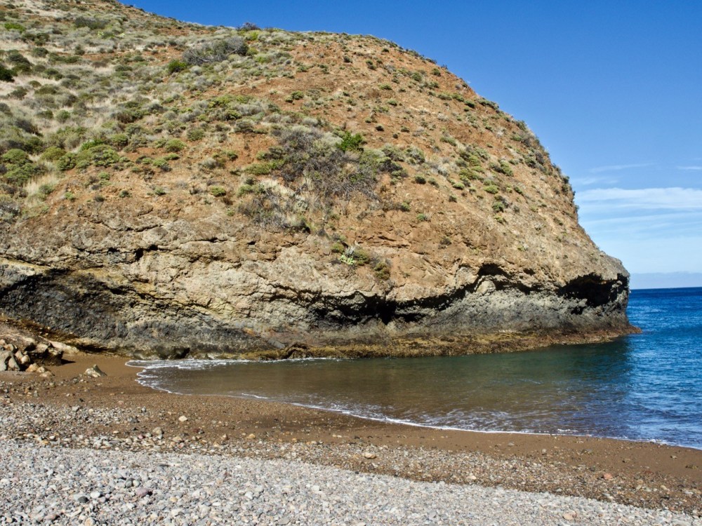 Rocky beach with a large cliff overlooking the ocean under a clear blue sky.
